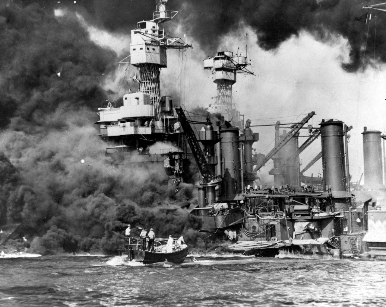 USS West Virginia burning and settling at her moorings, dense black smoke rising from the superstructure, a motor launch working close alongside her hull.