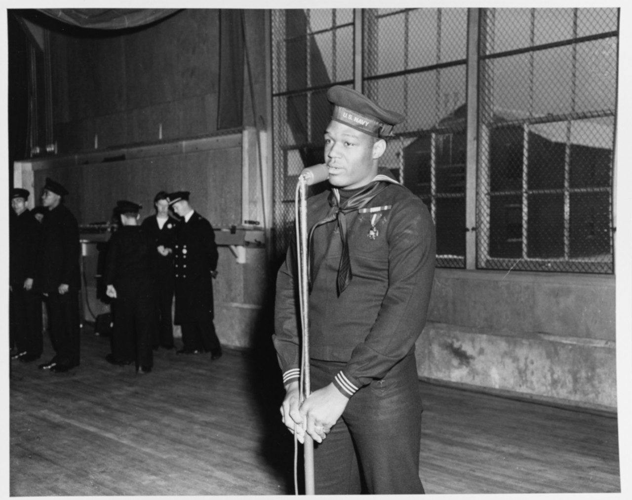 Doris Miller standing at a microphone in dark winter service dress, the Navy Cross on his chest, cap tally reading U.S. NAVY, with naval officers watching from the far side of a bare hall.