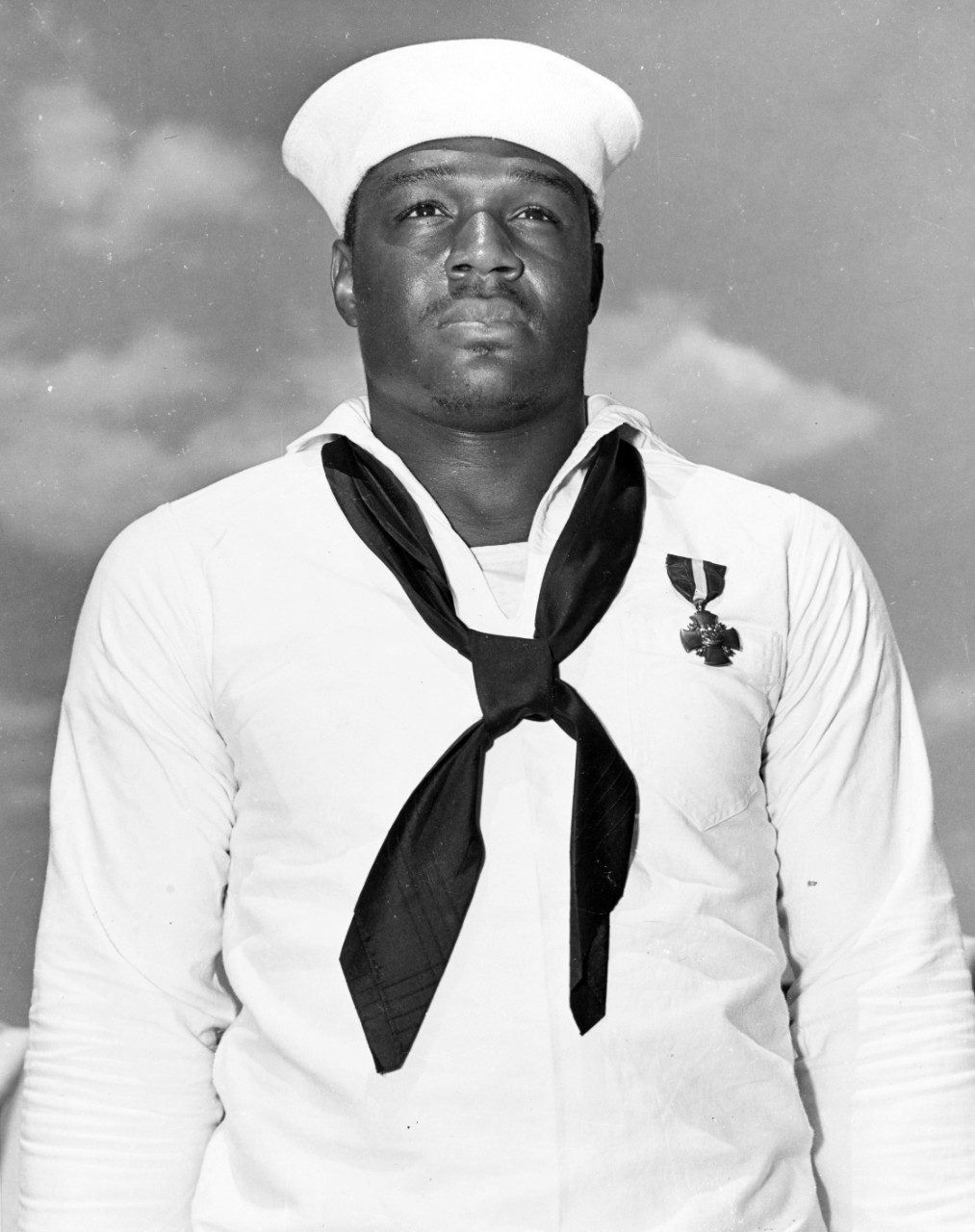 Studio portrait of Doris Miller in white summer service dress and white cap, black neckerchief, the Navy Cross pinned to his left breast, against a painted cloud backdrop.