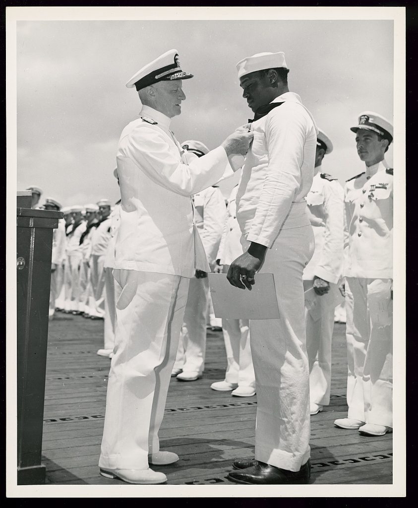 Admiral Chester Nimitz, in white service dress, pinning the Navy Cross to the jumper of Doris Miller, who stands at attention on a wooden deck holding his citation, ranks of officers behind them.